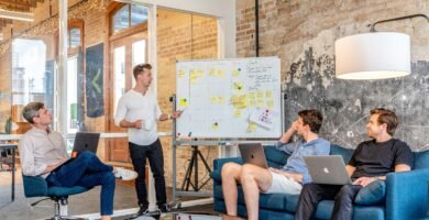 three men sitting while using laptops and watching man beside whiteboard